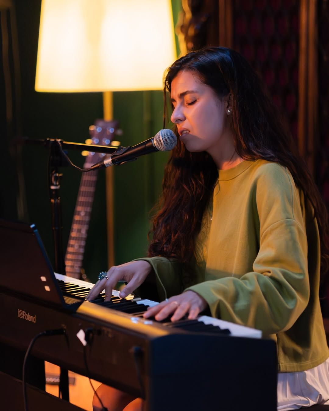 Olivia Mihet at the piano in a green sweater, lit by a warm lamp against a velvet backdrop
