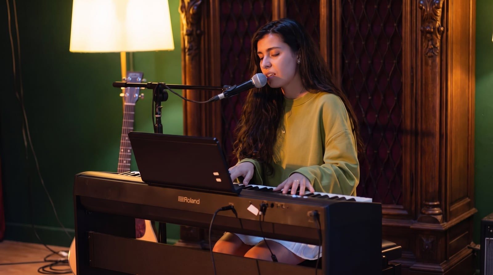 Olivia Mihet at the piano in a green sweater, lit by a warm lamp against a velvet backdrop