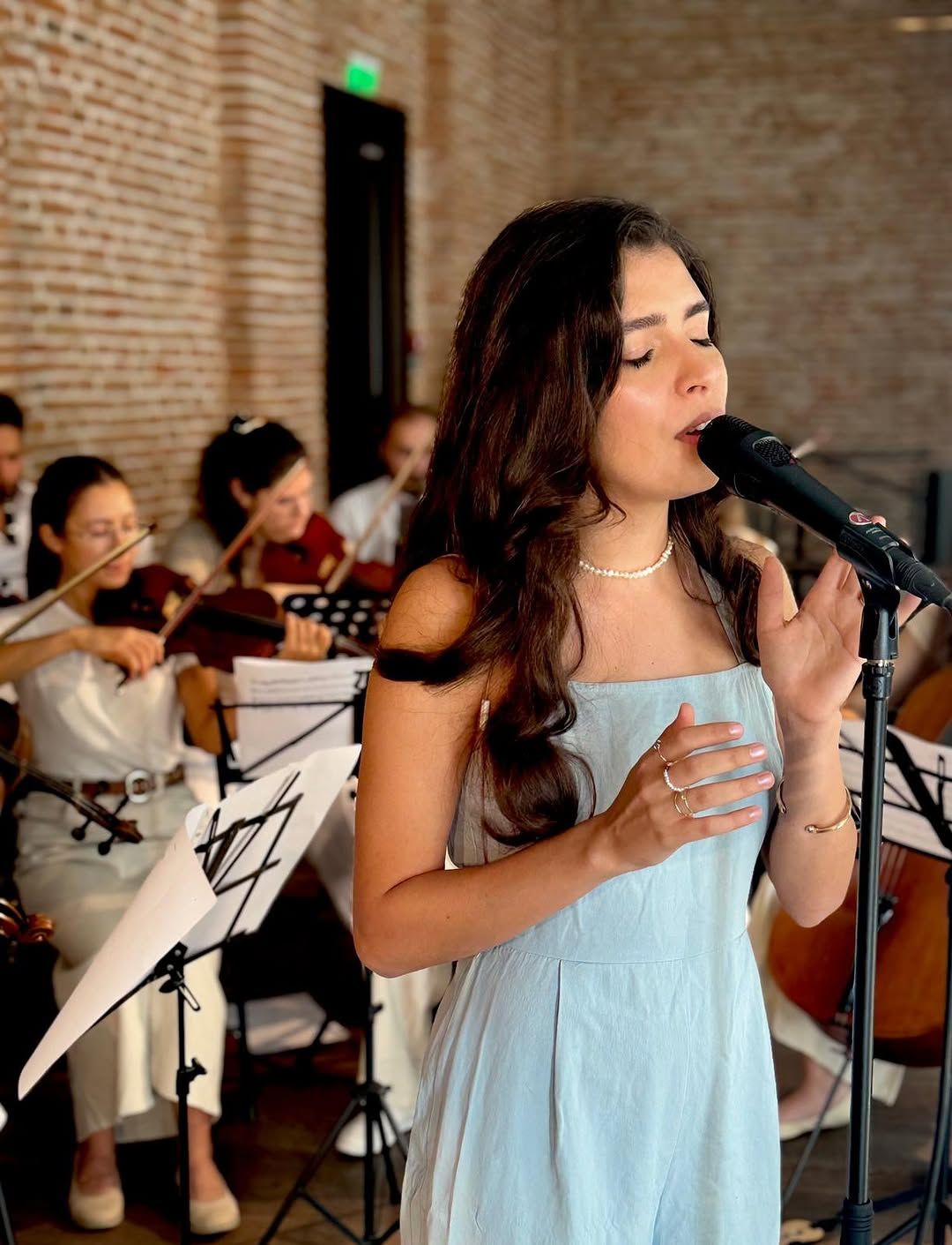 Close portrait of Olivia Mihet singing with eyes closed, violinists visible behind her at the same concert