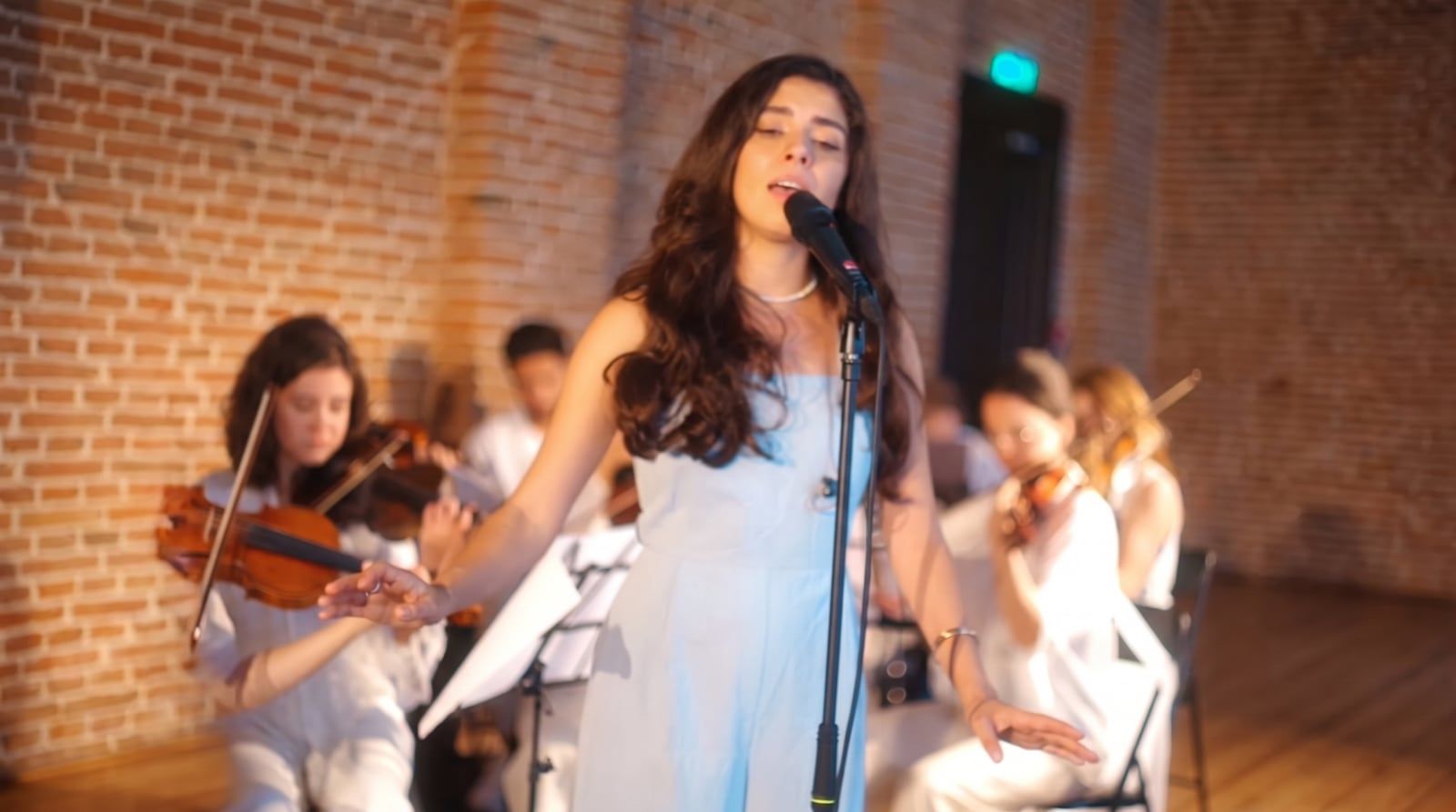 Olivia Mihet singing with arms open on stage with a string ensemble in a brick-walled hall