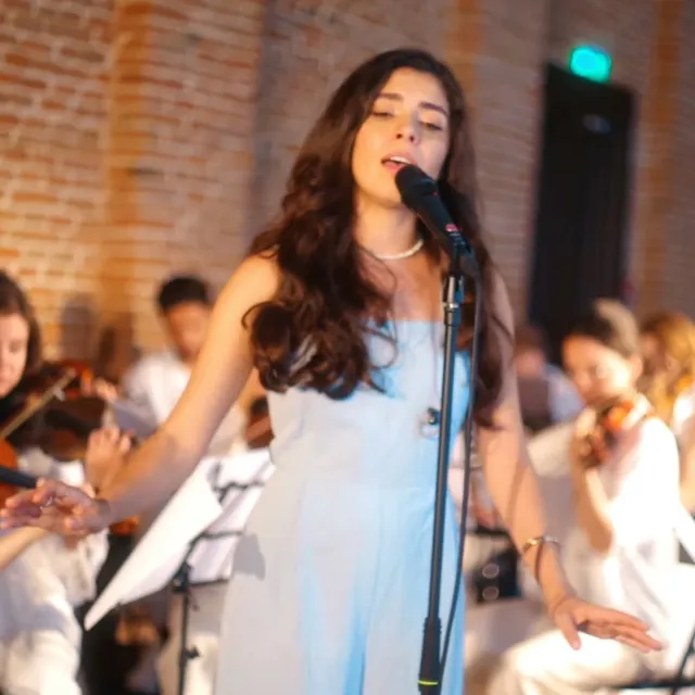 Olivia Mihet singing with arms open on stage with a string ensemble in a brick-walled hall