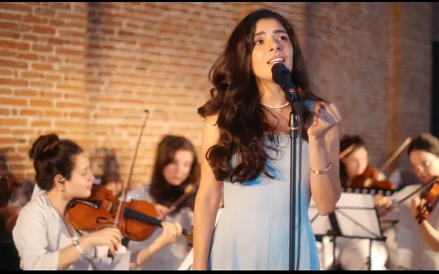 Olivia Mihet in a pale blue gown, singing with a string ensemble in a warmly lit, red-brick concert hall