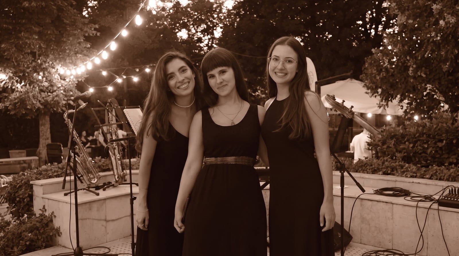 Olivia Mihet performing in a trio in black dresses on a garden stage strung with fairy lights, saxophone on a stand nearby