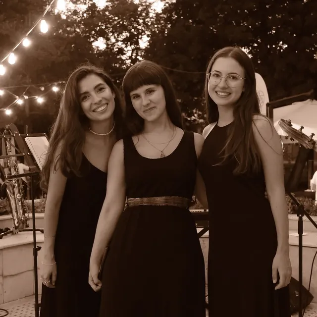 Olivia Mihet performing in a trio in black dresses on a garden stage strung with fairy lights, saxophone on a stand nearby