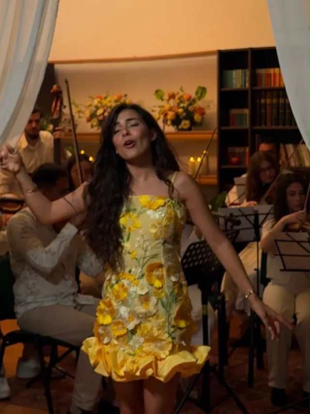 Olivia Mihet in a yellow floral dress singing in a library-style room with strings, sheer drapes and candlelight